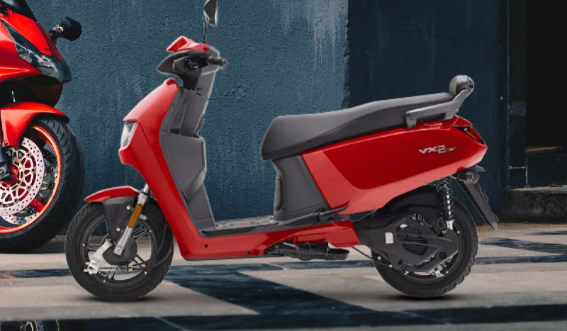 Create a featured image of Hero Vida VX2 electric scooter, in bright orange and white color, parked on a clean city street. Add icons of battery, mobile app, and 100km range. Show headline in top-left: ‘Hero Vida VX2 is Here’. Clean, youth-focused, eco-style layout. Avoid generic e-scooters — match actual VX2 model look.