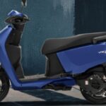 Create a featured image of Hero Vida VX2 electric scooter, in bright orange and white color, parked on a clean city street. Add icons of battery, mobile app, and 100km range. Show headline in top-left: ‘Hero Vida VX2 is Here’. Clean, youth-focused, eco-style layout. Avoid generic e-scooters — match actual VX2 model look.